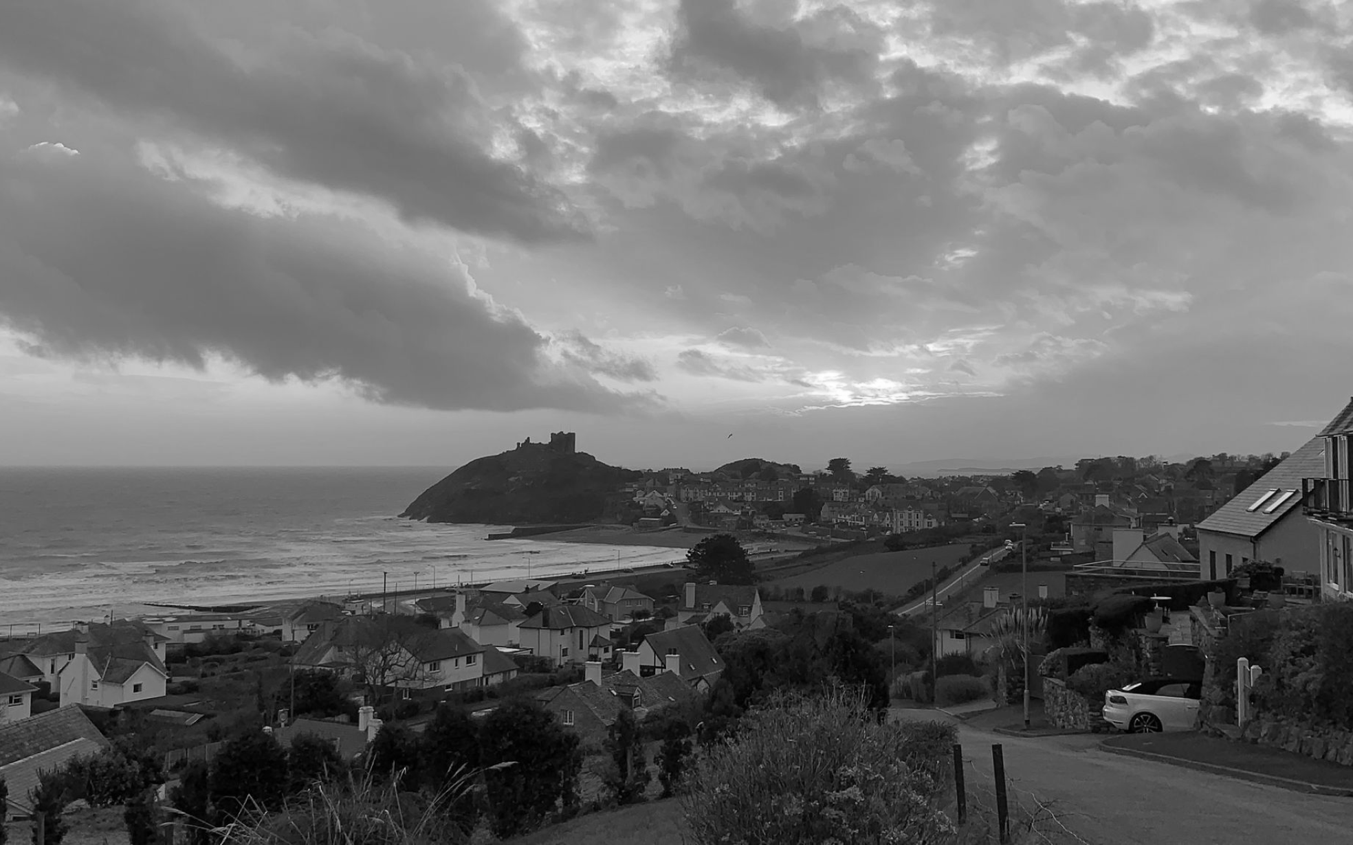 Vista panorâmica de uma cidade costeira ao entardecer, com casas no primeiro plano, um castelo sobre uma colina ao fundo e o oceano ao lado.